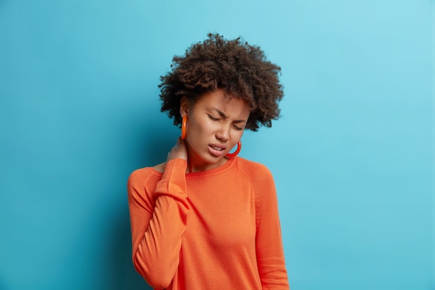 Infeliz joven sufre de dolor en el cuello se siente cansado masajes en el cuello siente incomodidad cierra los ojos viste un jersey naranja casual aislado sobre una pared azul