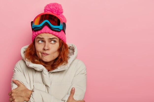 Foto gratuita infeliz joven tiembla de frío, frunce los labios, pasa mucho tiempo durante el clima helado, mira hacia el lado derecho, se encuentra sobre un fondo rosado