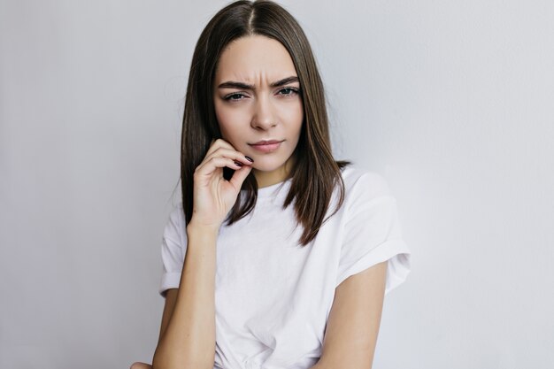 Infeliz mujer encantadora en camiseta posando. Chica europea de pelo oscura desagradable de pie delante de la pared blanca.