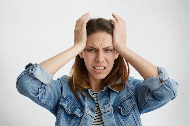 Infeliz mujer frustrada apretando los dientes, con mirada dolorosa, haciendo muecas, apretando las sienes, sufriendo de migraña o dolor de cabeza, sintiéndose estresado