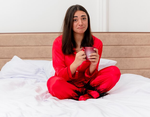 Infeliz mujer hermosa joven en pijama rojo sentado en la cama con una taza de café mirando a la cámara con expresión triste en el interior del dormitorio sobre fondo claro