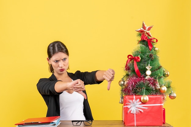 Infeliz mujer joven en traje haciendo gesto negativo cerca del árbol de Navidad decorado en la oficina en amarillo