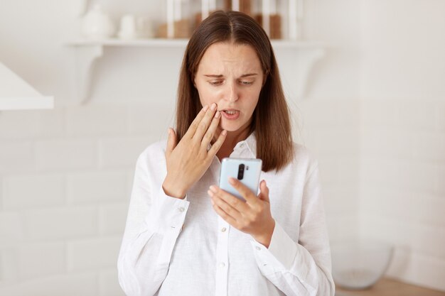 Infeliz mujer molesta de pie con teléfono inteligente en las manos, tener videollamadas, escuchar malas noticias, posando en la cocina en casa, mujer con camisa blanca de estilo casual.