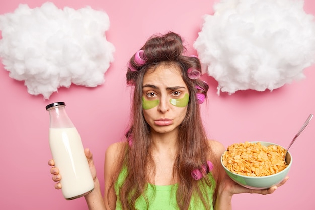 Infeliz mujer morena con rulos en la cabeza aplica colágeno verde almohadillas debajo de los ojos para tomar un desayuno saludable come copos de maíz con leche