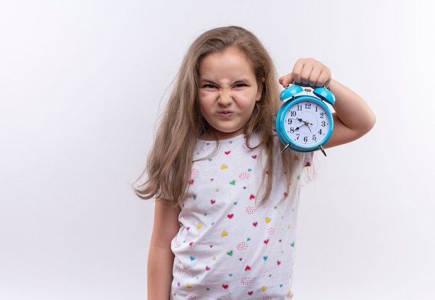 Infeliz niña de la escuela con camiseta blanca con reloj despertador en pared blanca aislada