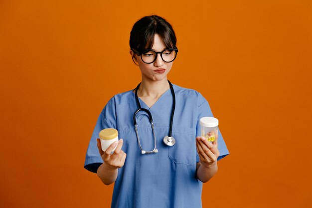 Infeliz sosteniendo envases de píldoras joven doctora vistiendo uniforme fith estetoscopio aislado sobre fondo naranja