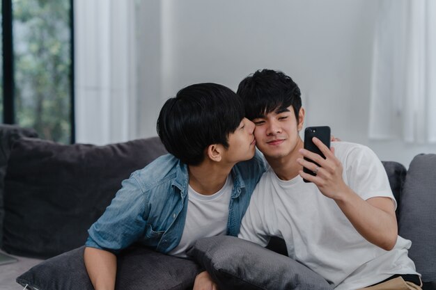 Influencia asiática Pareja gay vlog en casa. Los hombres LGBTQ asiáticos felices se divierten y se divierten usando la tecnología del teléfono móvil, graban videos de estilo de vida y suben videos a las redes sociales mientras descansan en el sofá de la sala de estar.