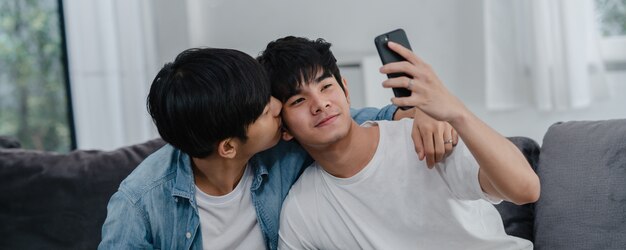 Influencia asiática Pareja gay vlog en casa. Los hombres LGBTQ asiáticos felices se divierten y se divierten usando la tecnología del teléfono móvil, graban videos de estilo de vida y suben videos a las redes sociales mientras descansan en el sofá de la sala de estar.