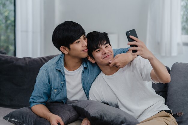 Influencia asiática Pareja gay vlog en casa. Los hombres LGBTQ asiáticos felices se divierten y se divierten usando la tecnología del teléfono móvil, graban videos de estilo de vida y suben videos a las redes sociales mientras descansan en el sofá de la sala de estar.