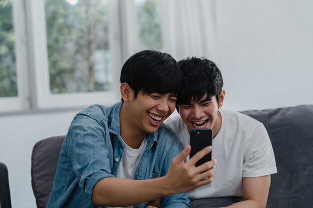 Influencia asiática Pareja gay vlog en casa. Los hombres LGBTQ asiáticos felices se divierten y se divierten usando la tecnología del teléfono móvil, graban videos de estilo de vida y suben videos a las redes sociales mientras descansan en el sofá de la sala de estar.