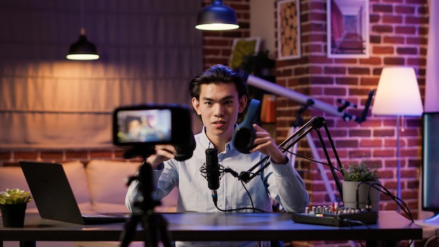 Influenciador asiático filmando revisión de auriculares en episodio de podcast, grabando video de recomendación en cámara. Programa de entrevistas de transmisión en vivo con producto de auriculares inalámbricos en las redes sociales.