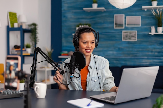 Influenciador de las redes sociales mirando una laptop y grabando videos. Mujer vlogger transmitiendo podcast en línea en un smartphone. Creador de contenido en estudio casero usando equipo de transmisión de audio.