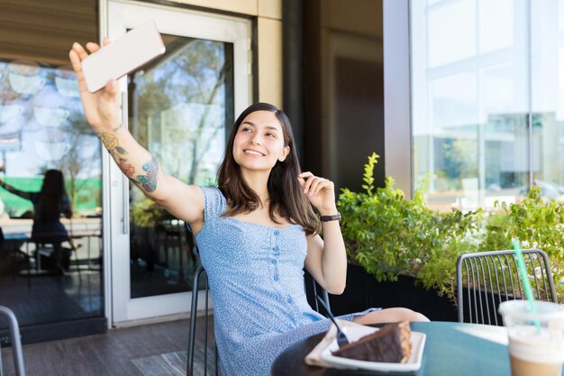 Influenciador sonriente tomando selfie mientras disfruta de pasteles y café en la cafetería en el centro comercial