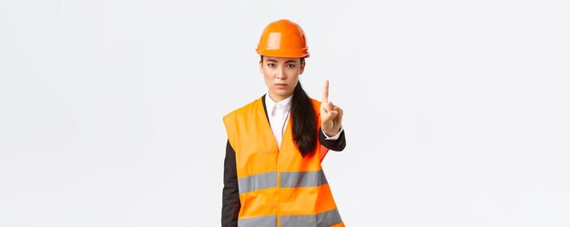 Ingeniera asiática de aspecto serio, confiada y disgustada, sacudiendo el dedo regañando a un trabajador de la construcción por actuar de forma peligrosa, prohibir y prohibir la acción en la zona de construcción de fondo blanco.