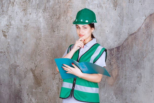 Foto gratuita ingeniera en casco verde sosteniendo una carpeta azul y parece confundida y pensativa.