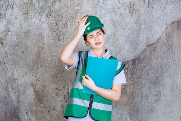 Ingeniera en casco verde sosteniendo una carpeta azul y poniendo mano a cabeza mientras está cansada.