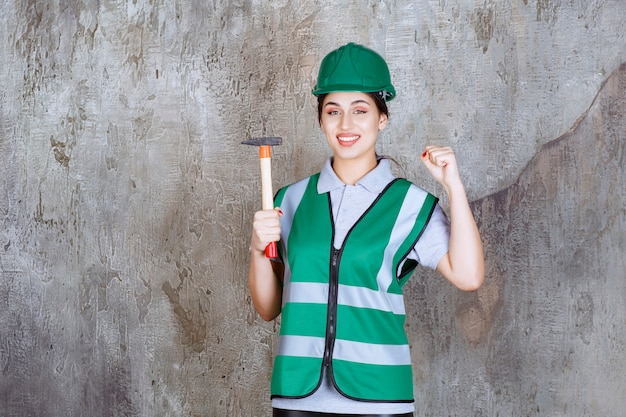Ingeniera en casco verde sosteniendo un hacha con mango de madera para un trabajo de reparación y mostrando los músculos de su brazo