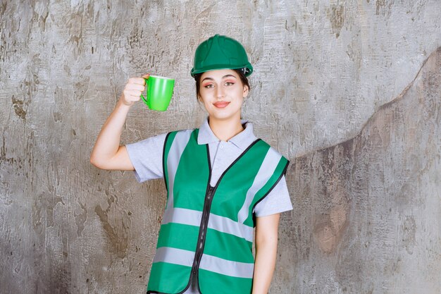 Ingeniera en casco verde sosteniendo una taza de café verde y sonriendo