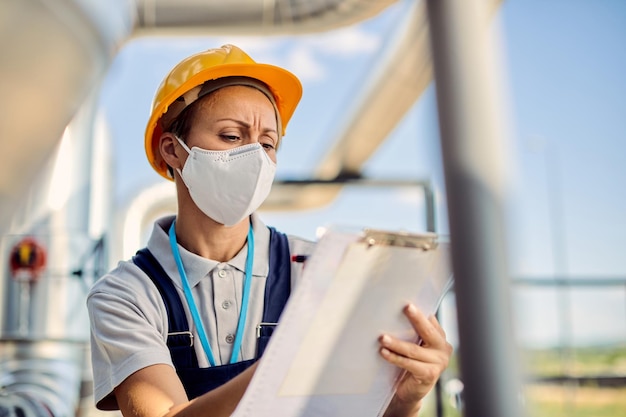 Ingeniera con mascarilla protectora escribiendo notas mientras inspecciona instalaciones industriales al aire libre