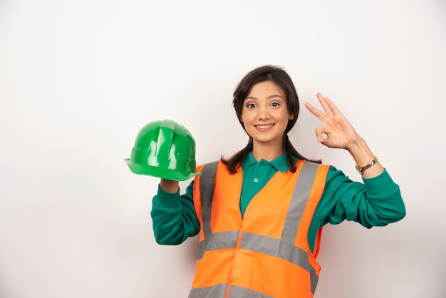 Foto gratuita ingeniera mostrando gesto ok y sosteniendo un casco sobre fondo blanco.