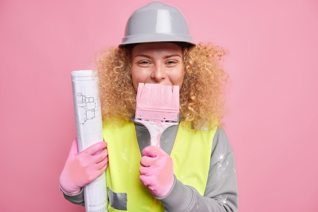 Ingeniera positiva con cabello rizado y tupido mantiene el pincel sobre la boca sostiene el modelo usa casco protector que refleja la chaqueta se divierte en el sitio de construcción. Inspector de construcción interior.