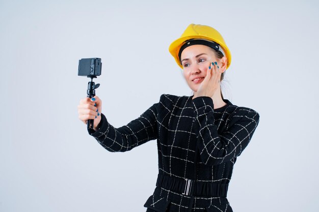 La ingeniera sonriente se está tomando una selfie con una mini cámara poniendo la mano en la mejilla sobre fondo blanco.