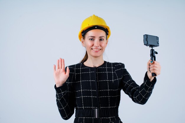 La ingeniera sonriente sostiene su mini cámara y muestra un gesto de saludo a la cámara con fondo blanco