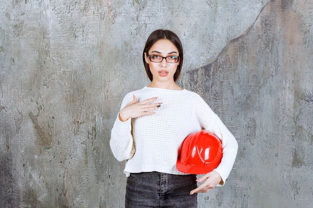 Ingeniera sosteniendo un casco rojo y apuntando a sí misma.