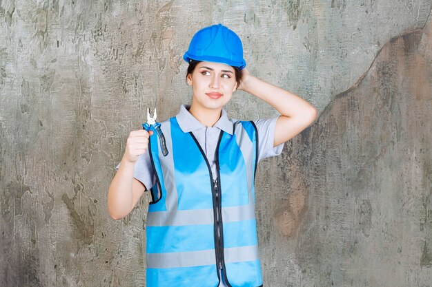 Ingeniera en uniforme azul y casco con alicates metálicos para reparación y se ve pensativa.