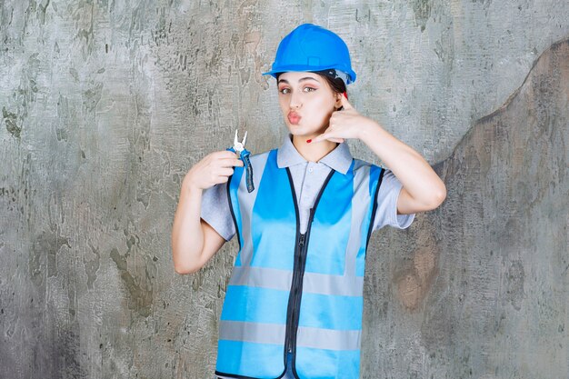 Ingeniera en uniforme azul y casco sosteniendo alicates metálicos para reparación y pidiendo una llamada