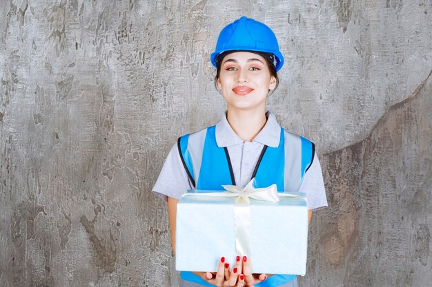 Ingeniera en uniforme azul y casco sosteniendo una caja de regalo azul.