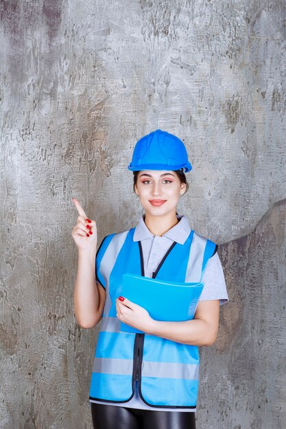 Ingeniera en uniforme azul y casco sosteniendo una carpeta de informe azul y apuntando a alguien alrededor