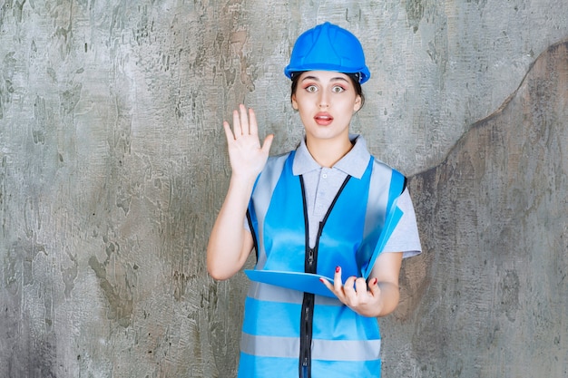 Ingeniera en uniforme azul y casco sosteniendo una carpeta de informe azul, leyendo y haciendo preguntas.