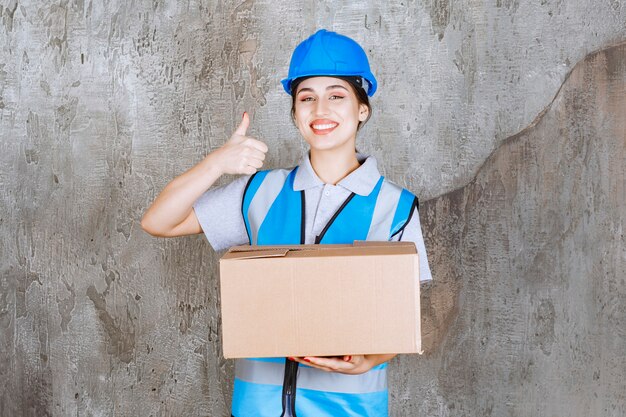 Ingeniera en uniforme azul y casco sosteniendo un paquete de cartón y mostrando un signo de mano positivo