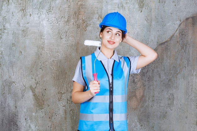 Ingeniera en uniforme azul y casco sosteniendo un rodillo de ajuste para pintar, pensar y planificar.