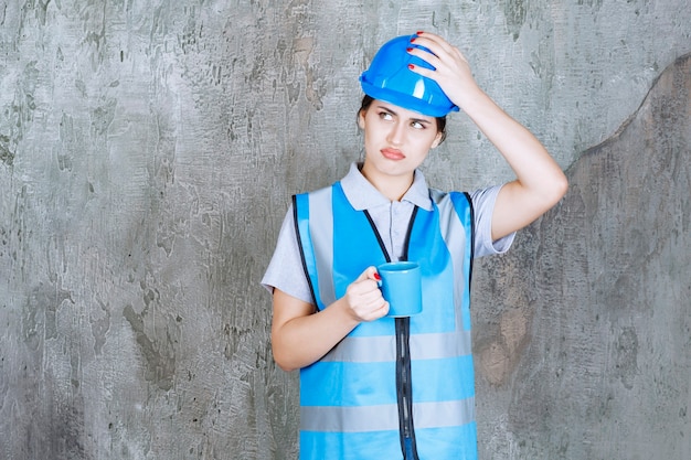 Ingeniera en uniforme azul y casco sosteniendo una taza de té azul y parece confundida y estresada.