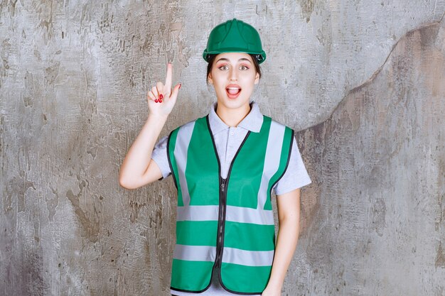 Ingeniera en uniforme verde y casco apuntando arriba con emociones