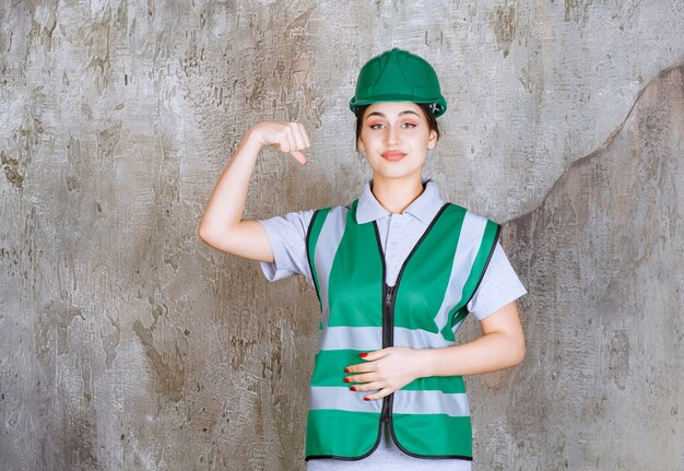 Ingeniera en uniforme verde y casco demostrando los músculos de su brazo.