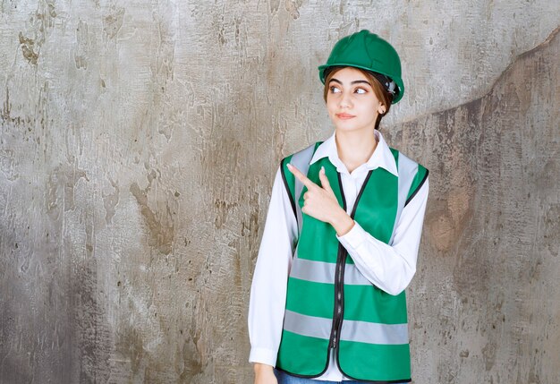 Ingeniera en uniforme verde y casco de pie en la pared de hormigón y apuntando hacia el lado izquierdo