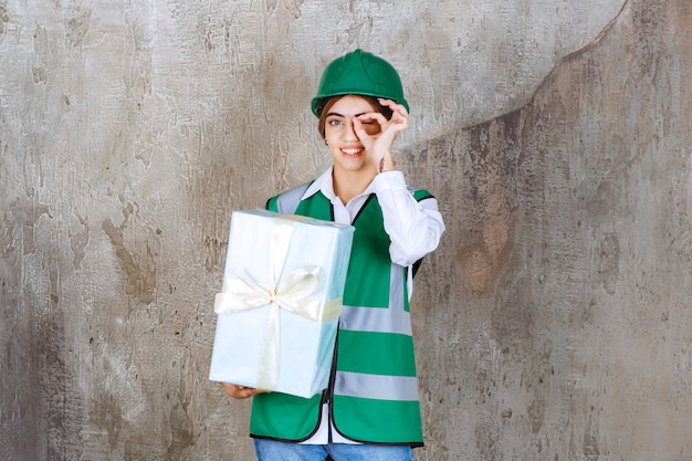 Ingeniera en uniforme verde y casco sosteniendo una caja de regalo azul y mostrando signo de disfrute.