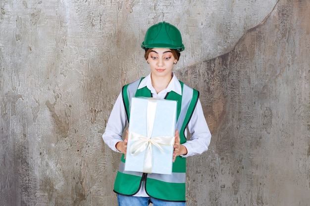 Ingeniera en uniforme verde y casco sosteniendo una caja de regalo azul.