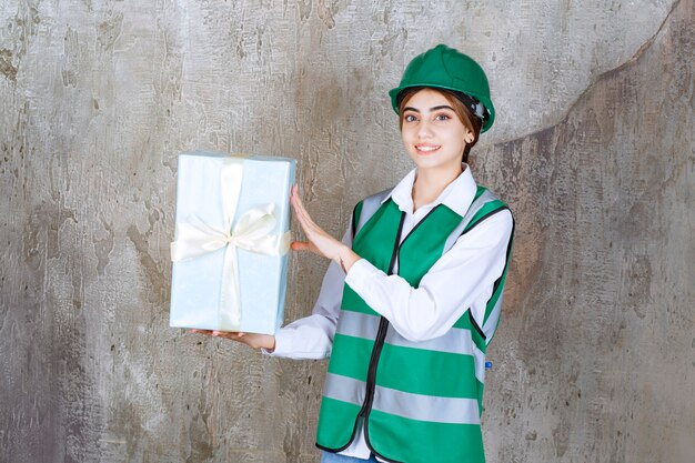 Ingeniera en uniforme verde y casco sosteniendo una caja de regalo azul.