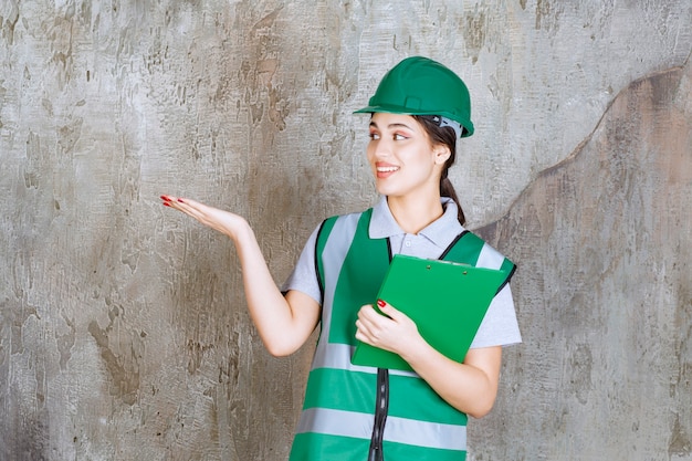 Ingeniera en uniforme verde y casco sosteniendo la carpeta del proyecto y apuntando a alguien.