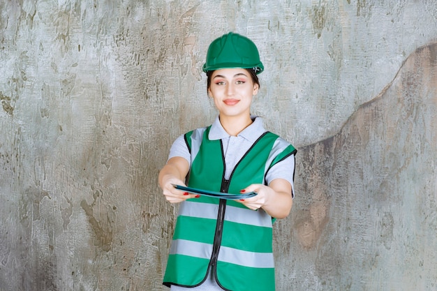 Foto gratuita ingeniera en uniforme verde y casco sosteniendo una carpeta de proyecto azul y ofreciéndola para verificación.