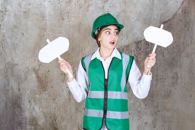 Foto gratuita ingeniera en uniforme verde y casco sosteniendo dos letreros en ambas manos y parece confundido