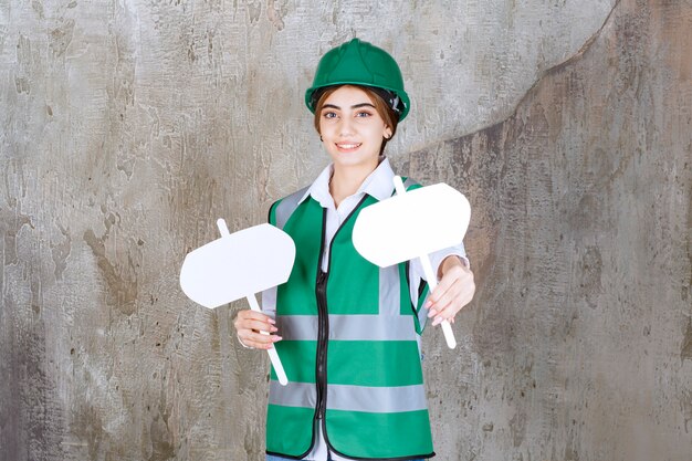 Ingeniera en uniforme verde y casco sosteniendo dos letreros en ambas manos.