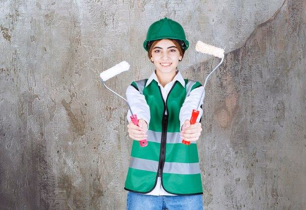 Ingeniera en uniforme verde y casco sosteniendo dos rodillos de ajuste para pintar con ambas manos y compartir con su colega.