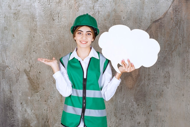 Ingeniera en uniforme verde y casco sosteniendo un tablero de información de forma de nube.