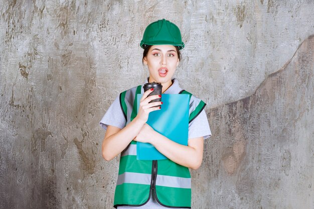 Ingeniera en uniforme verde y casco sosteniendo una taza de café negro y una carpeta de proyecto azul