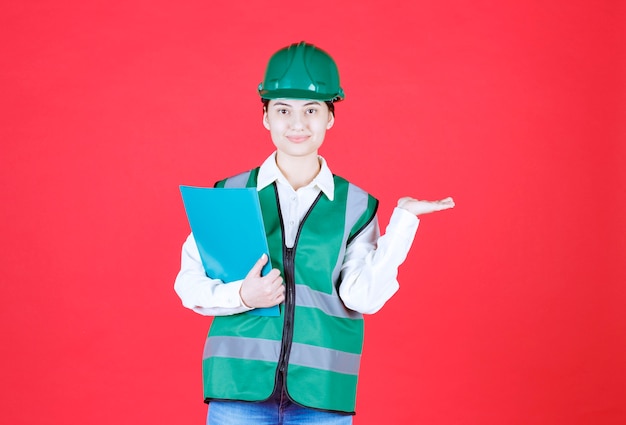 Ingeniera en uniforme verde sosteniendo una carpeta azul y llamando a su colega